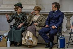 two men and a woman sitting on a bench in front of a building, talking