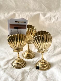 three gold shell shaped candlesticks sitting on a white sheet with a magazine in the background