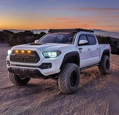 a white toyota pickup truck parked on top of a sandy hill at sunset with the sun setting in the background