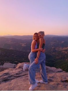 two women hugging each other while standing on top of a mountain at sunset with mountains in the background