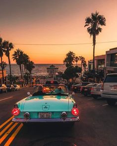 a blue car driving down a street next to the ocean at sunset with palm trees