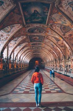 a woman walking down a long hallway with paintings on the walls and ceiling above her