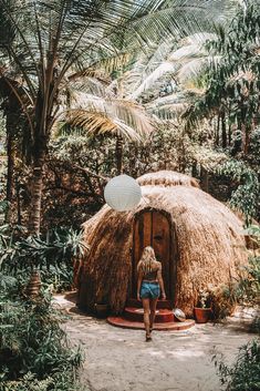 a woman standing in front of a straw hut surrounded by palm trees and foliage, with a white paper lantern hanging from the ceiling