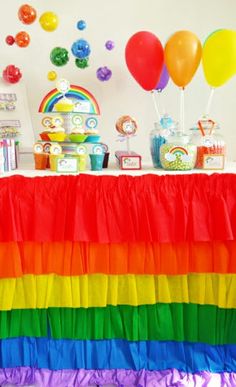 a rainbow themed party with balloons and streamers on the wall, table cloth draped over it