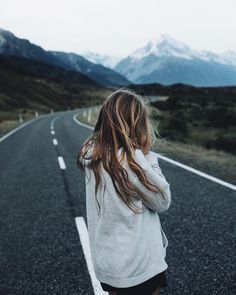 a woman standing on the side of an empty road with mountains in the background and her hair blowing in the wind