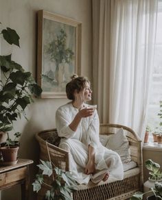 a woman sitting on top of a wicker couch next to a window with potted plants