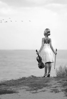 a woman standing on top of a sandy beach next to the ocean holding a violin