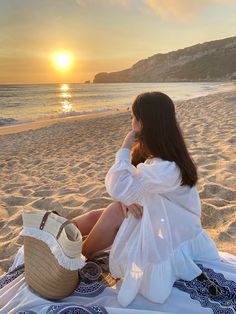 a woman sitting on top of a beach next to the ocean with a straw bag