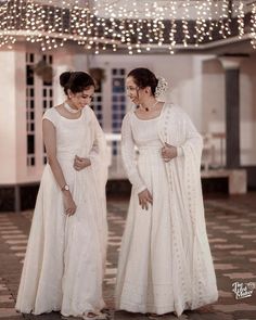 two women in white dresses standing next to each other on a tiled floor with lights overhead