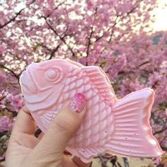 a hand holding up a pink fish shaped object in front of a tree with pink flowers