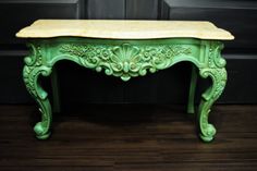 a green table with a wooden top and carvings on the legs, sitting on a hard wood floor