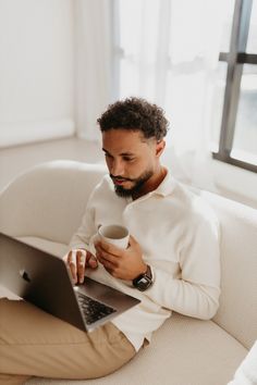 a man sitting on a couch holding a cup and looking at his laptop computer screen