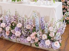 the table is covered with purple flowers and white vases filled with greenery on it