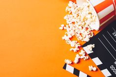 popcorn spilled out of a bucket next to a clapper and movie ticket on an orange background