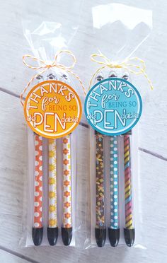 two pens in plastic packaging on top of a wooden table next to another pen with an orange, blue and yellow design