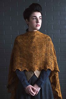 a woman standing in front of a brick wall wearing a brown shawl and jeans