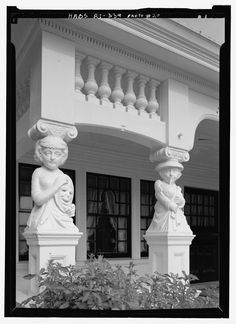 black and white photograph of two statues in front of a building with plants growing outside