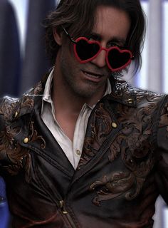 a man with heart shaped sunglasses on his face looking at the camera while wearing a leather jacket and white shirt