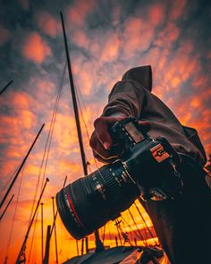 a person holding a camera in front of a sunset with sailboats on the water