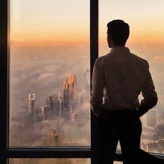 a man standing in front of a window looking out at the city below and skyscrapers