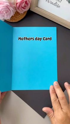 two hands holding a blue card with the words mothers day card on it and pink roses