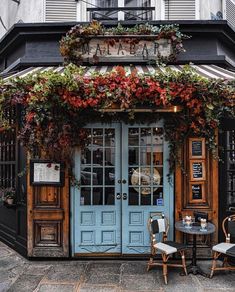 an outdoor cafe with blue doors and flowers on the outside wall, two chairs at a table in front of it