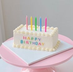 a birthday cake with lit candles on it sitting on a pink tablecloth in front of a white wall