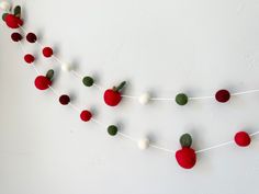 red and green felt apple garland hanging on wall with white string, pom - poms