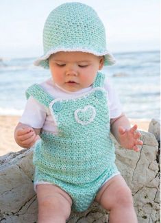 a baby wearing a green crochet outfit and hat sitting on a rock near the ocean