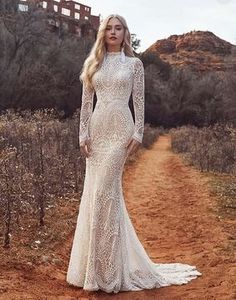 a woman standing on a dirt road wearing a long sleeved wedding dress with an open back