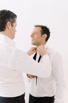 a man helping another man put on his tie in front of the camera while wearing a white shirt and black pants