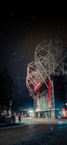 the stadium is lit up at night with snow falling on it and people walking around