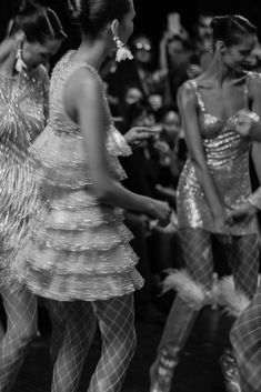 three women in shiny dresses standing next to each other with feathers on their legs and one woman looking at her phone