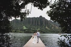 a bride and groom are walking on a dock near the water with mountains in the background