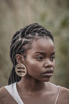 a woman with dreadlocks standing in front of some bushes and trees, looking off to the side