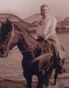 a woman riding on the back of a brown horse next to a body of water