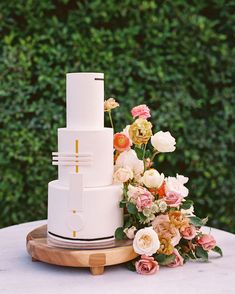 a three tiered cake with flowers and greenery behind it on a wooden platter