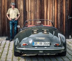 a man standing next to an old fashioned sports car in front of a wooden fence