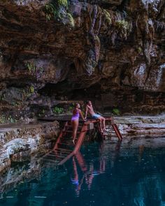 two women in bikinis are sitting at the edge of a pool near a cliff