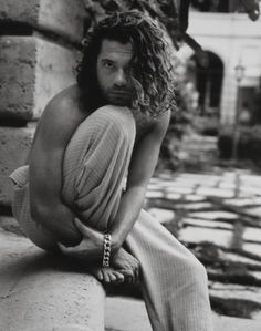 a man with long hair sitting on the ground next to a brick wall and stairs