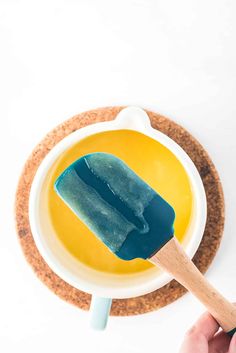 a hand holding a blue ice cream on top of a yellow bowl with a wooden spoon