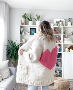 a woman standing in front of a white bookcase holding a pink heart sweater over her shoulders