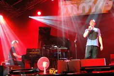 two men standing on stage with microphones in front of them and music equipment behind them