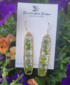 a pair of earrings with green flowers on them sitting in front of some purple flowers