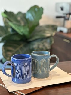 two coffee mugs sitting on top of a table next to a potted plant