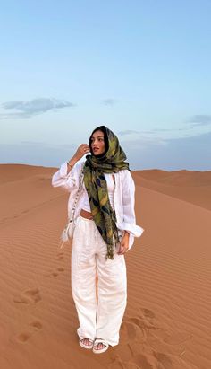 a woman standing in the middle of a desert wearing white pants and a green scarf