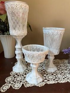 three white vases sitting on top of a doily covered table next to potted plants