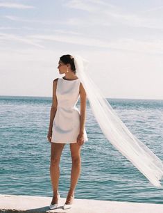 a woman in a short white dress and veil standing on the edge of a pier