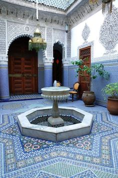 a fountain in the middle of a courtyard with potted plants and doors behind it