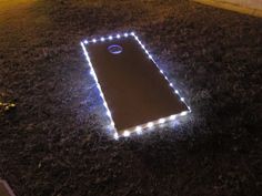 an illuminated cornhole in the middle of a yard with grass and lights around it